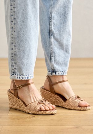 Fødder iført lysebrune flettede kilesandaler med ankelremme og lyseblå denimjeans på et trægulv.