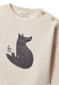 Chemise beige en coton à manches longues avec un tissu texturé, présentant une application de renard gris foncé à l'avant et des boutons en bois à l'épaule.