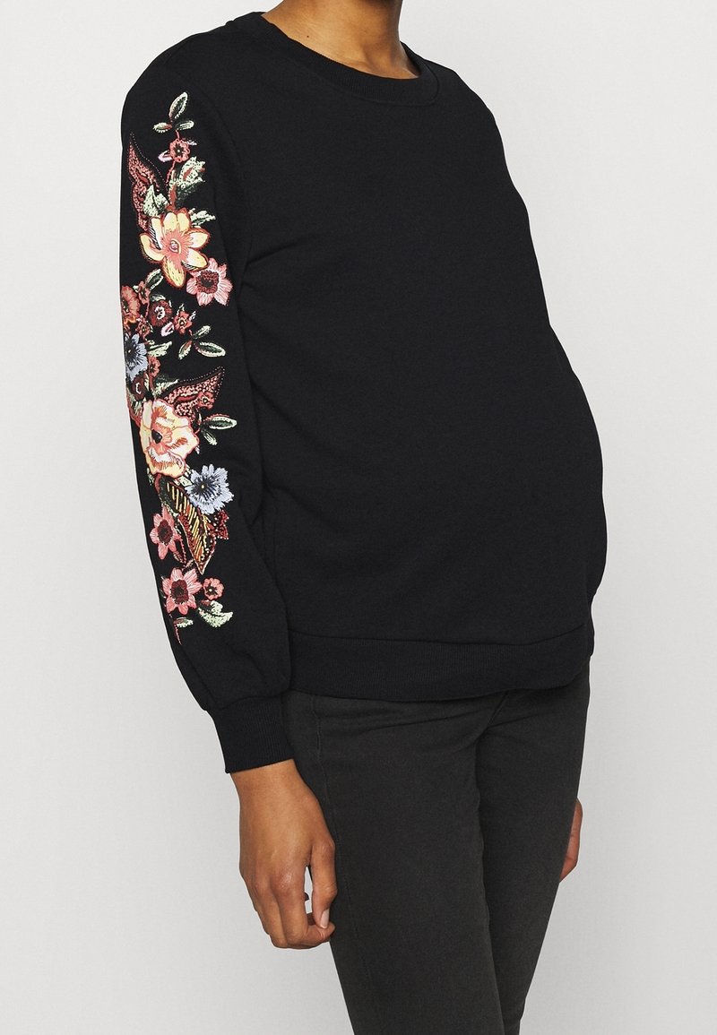 Personne enceinte portant un pull noir à manches longues avec une broderie florale colorée sur la manche droite et un pantalon noir.