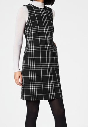 Kvinde iført en ærmeløs sort kjole med hvidt ternet mønster over en hvid langærmet top og sorte strømpebukser, stående mod en hvid baggrund.