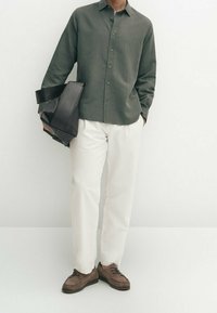 Donkergroene blouse met lange mouwen, witte broek, bruine schoenen; houdt een zwarte draagtas vast. De stof is textuurvol en het ontwerp is casual.