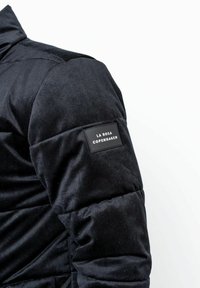 Zwarte gewatteerde jas met een zachte ribbelstructuur. Voorzien van een zwart merklabel op de linker mouw. Hoog col ontwerp.