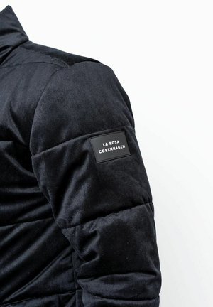 Sort quiltet jakke med en blød ribbet tekstur. Har en mærket sort label på venstre ærme. Design med høj krave.