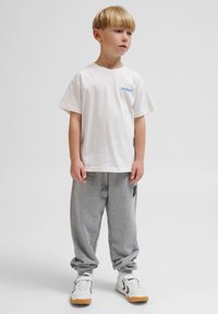 T-shirt blanche à manches courtes avec un logo bleu sur la poitrine gauche, associée à un pantalon de jogging gris clair et des baskets blanches avec des semelles brunes.