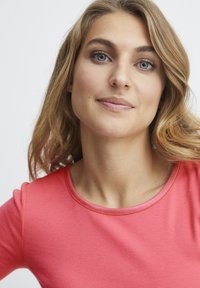 Korálová košile s žebrovaným vzorem a kulatým výstřihem; má jemnou texturu a obepínající střih. Vlasy jsou vlnité a volně spadají na ramena.