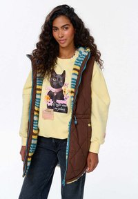 Pruunist quiltitud vestist sinise ja kollase triibulise äärega, kantud kollase sviitri peal, millel on musta kassi graafika ja tekst. Tõmblukuga teksapüksid.