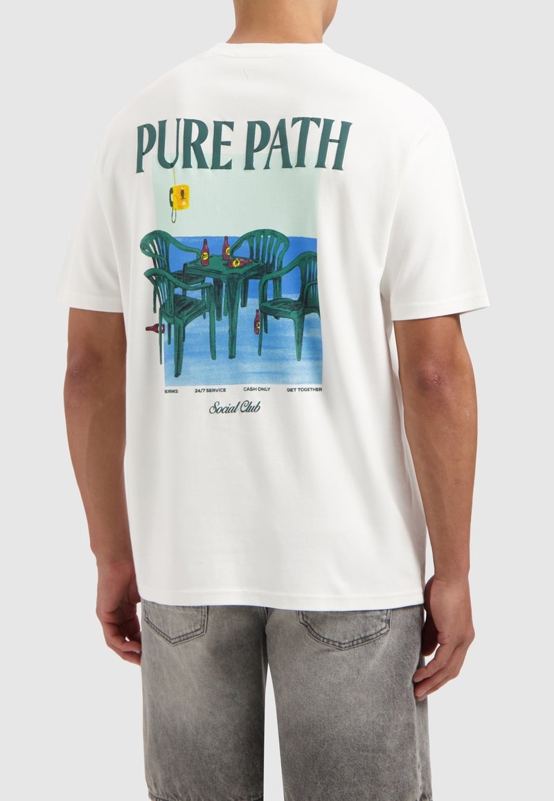 Witte t-shirt met een kleurrafische afbeelding op de achterkant, die groene stoelen, een tafel en flessen tegen een blauwe achtergrond afbeeldt.
