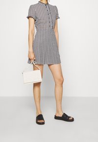 Schwarz-weißer Karokleid mit kurzen Ärmeln, Knopfleiste und ausgestelltem Rock. Kombiniert mit einer kleinen weißen Handtasche und schwarzen Sandalen.