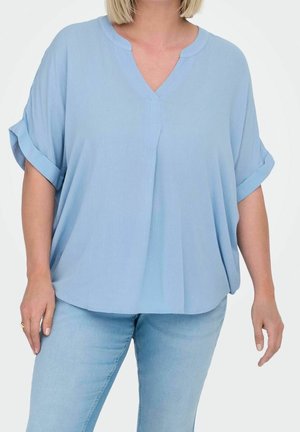 Vrouw draagt een lichtblauw V-halsblouse met opgerolde mouwen en lichtblauwe spijkerbroek tegen een witte achtergrond.