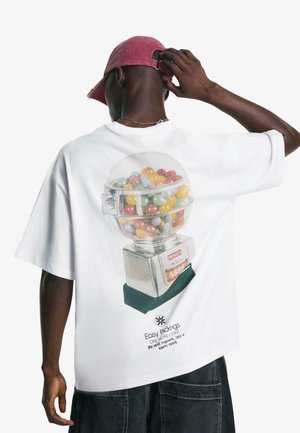 Hvid oversized t-shirt med et farverigt gumball-maskine grafisk design på ryggen. Stoffet er blødt; designet inkluderer tekst og detaljer.