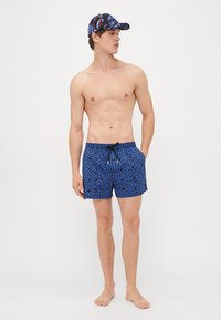 Ung man som bär blå mönstrade badshorts och en svart och blå mönstrad mössa, stående barfota mot en enkel bakgrund.