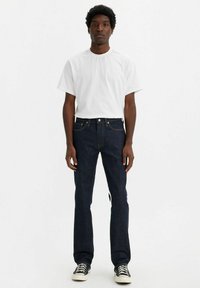 Camiseta de algodón blanca combinada con jeans de mezclilla azul oscuro. Los jeans tienen un corte recto y costuras claras en los bolsillos. Zapatillas negras.