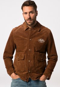 Veste en daim marron avec un devant à boutons, un col pointu et deux poches avant. Présente un patch logo sur la poitrine avec une texture douce.