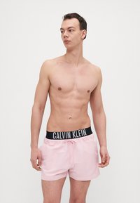 Rosafarbene Badeshorts mit einem schwarzen elastischen Bund, auf dem das "Calvin Klein"-Logo prangt. Glatte Textur und Design in Oberschenkel-Länge.