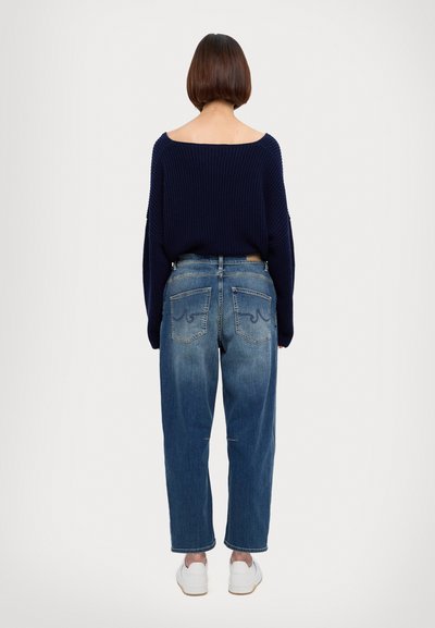 Maglione navy a coste con spalle scoperte abbinato a jeans azzurri larghi. I jeans presentano una texture sbiadita e due tasche posteriori con dettagli di cucitura. Sneakers bianche.