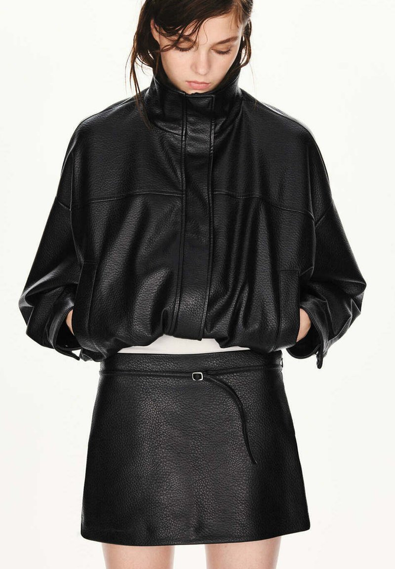 Jeune femme portant une veste en cuir noire texturée oversize et une mini-jupe assortie, regardant vers le bas avec les mains dans les poches de la veste.