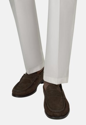 Mocassins en daim marron portés avec un pantalon blanc sur mesure, montrant des coutures détaillées et des semelles en caoutchouc sur une personne debout.