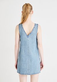 Denimmekko V-muotoisella selkäosalla ja ilman hihoja. Vaalea sininen väri, suora leikkaus ja näkyvät ompeleet reunojen ympärillä. Keskijalkaan ulottuva pituus.