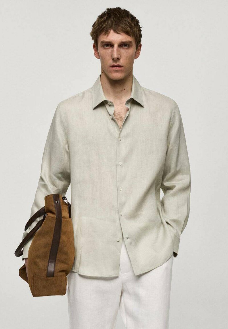 Camicia di lino chiara con maniche lunghe e chiusura a bottoni, abbinata a una borsa in suede marrone. Il modello indossa pantaloni color crema.