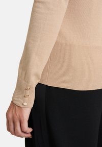 Poignet de pull côtelé beige avec trois boutons dorés associé à un pantalon noir, main détendue sur le côté.