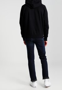 Sudadera con capucha negra de corte relajado, combinada con jeans azul oscuro y zapatillas blancas. Cuenta con un bolsillo tipo canguro y puños acanalados.