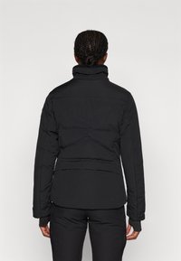 Melna pūkainā jaka ar augstu apkakli, pildītu struktūru un gludu tekstūru. Iespējas ietver izgrieztu jostasvietu un garas piedurknes ar elastīgām aprocēm.