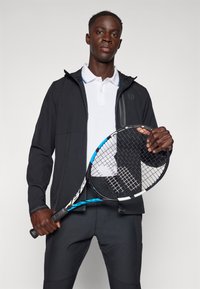 Must debel, mille tõmbluku ja valge polo särk helesiniste aktsentidega, ning mustad püksid. Hoides sinistest aktsentidest tennisereketit.