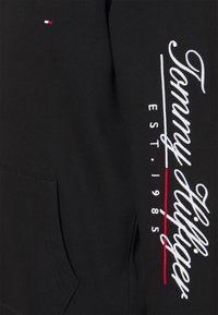 Sudadera negra con un bolsillo frontal. Presenta el texto bordado "Tommy Hilfiger" en blanco y un pequeño logo en rojo y azul en el pecho.