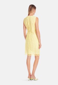 Gelbes Spitzenkleid mit einem figurbetonten Design, ärmellosem Schnitt und gewelltem Saum. Der Stoff weist ein strukturiertes Blumenmuster auf.