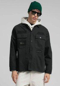 Chaqueta negra con botones y cuatro bolsillos delanteros, con etiquetas bordadas. Se lleva sobre una sudadera con capucha gris. Los accesorios incluyen un gorro verde y gafas de sol.