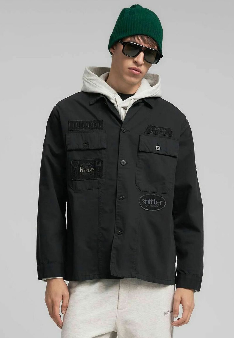 Chaqueta negra con botones y cuatro bolsillos delanteros, con etiquetas bordadas. Se lleva sobre una sudadera con capucha gris. Los accesorios incluyen un gorro verde y gafas de sol.