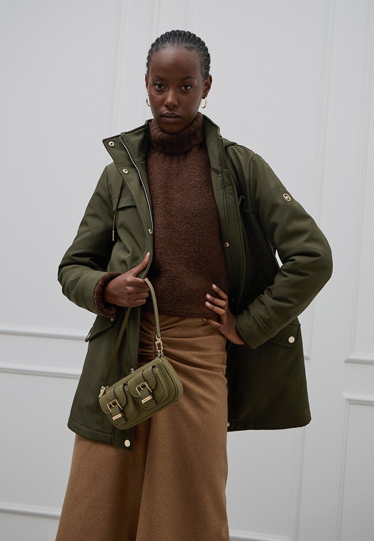 Giacca parka impermeabile verde oliva con chiusura a zip e bottoni a pressione, abbinata a un maglione marrone a maglia e a una gonna color sabbia, mentre tiene una borsa oliva.
