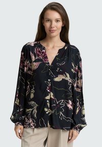Kvinna klädd i en lös svart blus med rosa, krämfärgade och gröna blommönster samt beige byxor, stående mot en ljusgrå bakgrund.