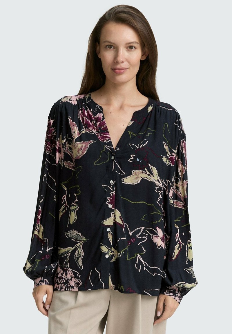 Kvinna klädd i en lös svart blus med rosa, krämfärgade och gröna blommönster samt beige byxor, stående mot en ljusgrå bakgrund.