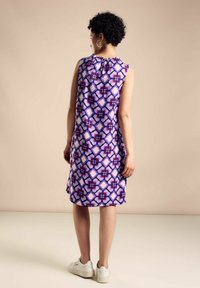 Lila ärmelloses Kleid mit geometrischem Muster, leichtem Stoff, gerafftem Ausschnitt und geradem Schnitt, kombiniert mit weißen Turnschuhen.