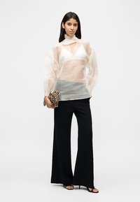 Γυναίκα που φοράει διάφανο λευκό πουκάμισο πάνω από λευκό μπραλέτο, μαύρο παντελόνι με φαρδιά πόδια, μαύρες γόβες και κρατάει μια τσάντα φάκελο με μοτίβο λεοπάρ.