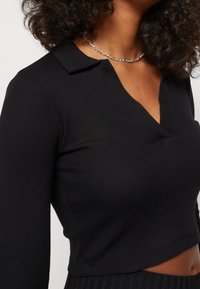 Schwarzes, geripptes Oberteil mit langen Ärmeln, V-Ausschnitt und Kragen. Der kurze Schnitt betont die Textur. Kombiniert mit einer silbernen Halskette.