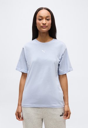 Γυναίκα με μακριά ίσια μαλλιά φοράει ανοιχτό μπλε μπλουζάκι Puma και ανοιχτό γκρι φόρμες Puma, στέκεται μπροστά σε απλό φόντο.