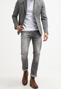 Grå blazer över en vit polo, ljusgrå jeans och bruna snörkängor. Detaljer inkluderar uppvikta ärmar och en ficka på blazern.