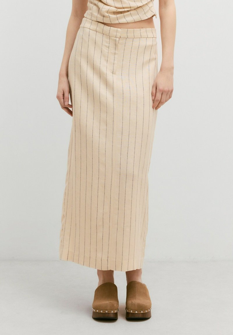 Beige linnen rok met verticale blauwe strepen, een aansluitende tailleband en een rechte silhoutte. Gecombineerd met bruine klompen versierd met metallic accenten.