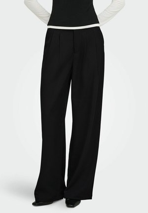 Personne portant un pantalon noir à jambes larges, des chaussures noires et un haut à manches longues noir et blanc, debout les mains dans le dos.