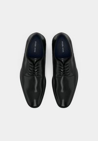 O pereche de pantofi de dress din piele neagră lustruită cu șireturi, văzuți de sus pe un fundal alb simplu.