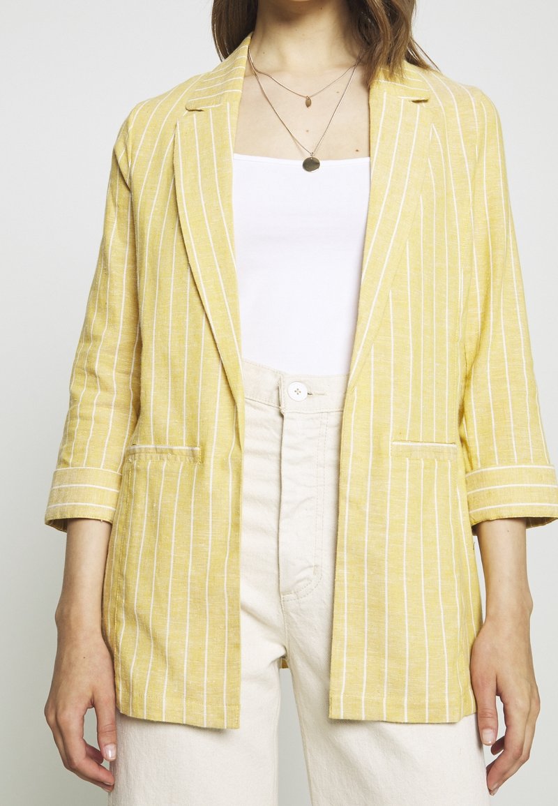 Żółty marynarka w paski o lnianej fakturze, z klapą z wycięciem, dwoma przednimi kieszeniami i rękawami do łokcia. W zestawie z białym topem.