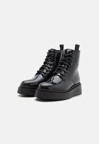 Botines de charol negro con diseño de cordones, punta redonda y suela gruesa de plataforma negra. Presentan tirador en la parte posterior.