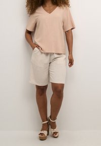 Top corto a V in rosa chiaro abbinato a pantaloni corti in lino beige. La modella indossa sandali con plateau spessi dal design annodato.