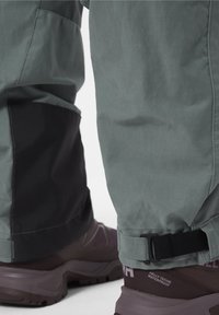 Voděodolné kalhoty v tlumené zelené barvě s černými detaily, s nastavitelnými manžetami a vyztuženými prvky, doplněné o tmavé, robustní boty.