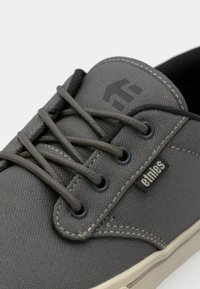 Sneaker in tela grigia con lacci rotondi, occhielli in metallo e logo nero sulla linguetta. Include cuciture a contrasto e una superficie testurizzata.