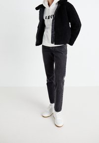 Person wearing a black fleece jacket, white hoodie with "LEVI'S" text, black jeans, white sneakers, and matching white socks against a plain background.