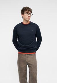 Jeune homme aux cheveux courts et bouclés, portant des lunettes, un sweat-shirt bleu marine par-dessus une chemise orange et un pantalon marron, les mains derrière le dos, regardant sur le côté.