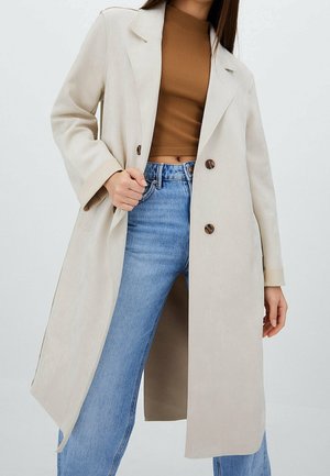 Mujer lleva un abrigo largo beige sobre un top corto marrón y vaqueros azul claro de talle alto, de pie con una mano sosteniendo el frente del abrigo.